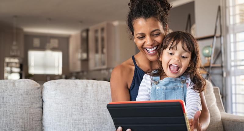 A mother and daughter smile at the laptop they're holding while sitting on a couch