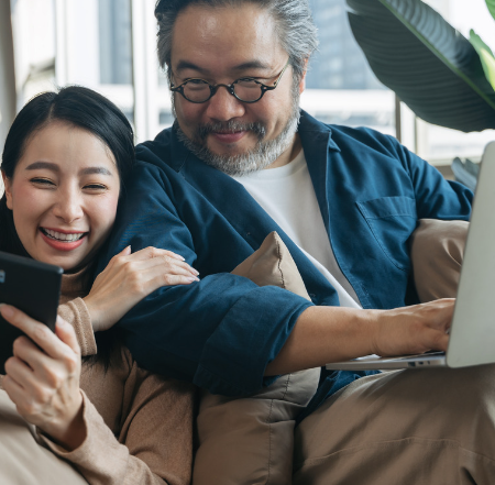 A smiling woman shows her phone to a smiling older man while he types on a laptop